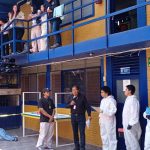 Estudiantes y docentes de criminología realizan una práctica de procesamiento de escena del crimen en un patio universitario. Se observa a personas en trajes de bioseguridad blancos frente a un área acordonada con cinta amarilla, mientras otros observan desde las escaleras y un pasillo superior.