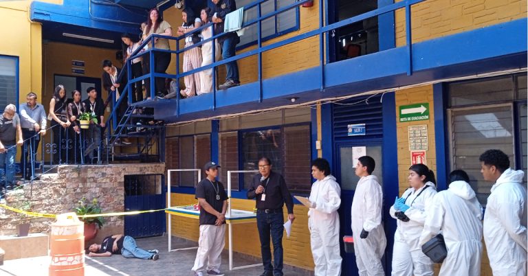 Estudiantes y docentes de criminología realizan una práctica de procesamiento de escena del crimen en un patio universitario. Se observa a personas en trajes de bioseguridad blancos frente a un área acordonada con cinta amarilla, mientras otros observan desde las escaleras y un pasillo superior.